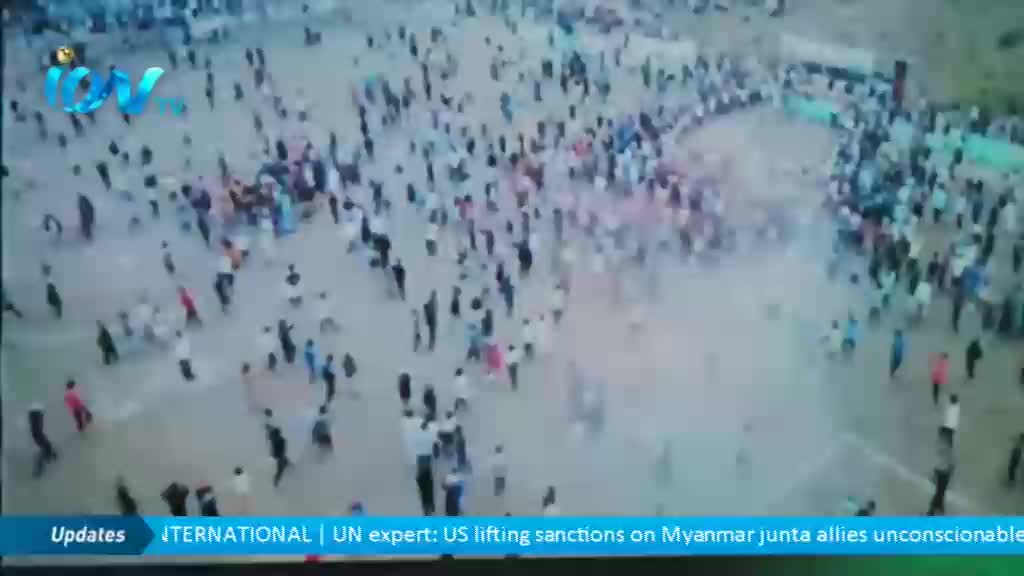 A large crowd of people disperses across a dusty expanse.  From the angle, it looks like the scene is being broadcast on ITV News.
