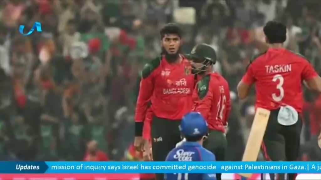 Three cricket players in red jerseys are standing on the field, while one, "Taskin" with the number 3 on his back, is walking away with a bat. A young boy in a blue helmet is facing them.
