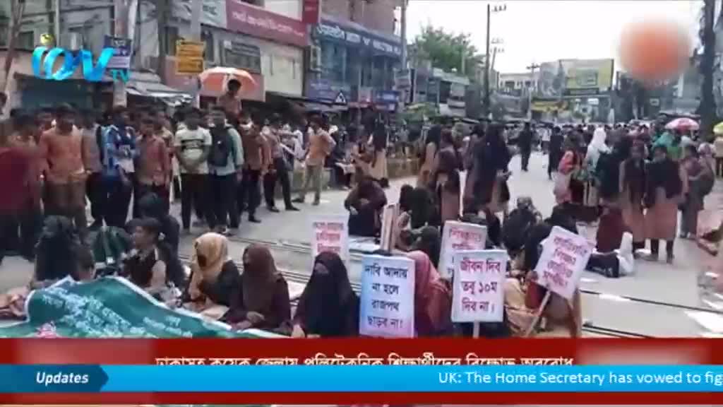People are gathered in the street, some holding signs with writing on them. A group is sitting on the ground, while others stand and walk in the background.
