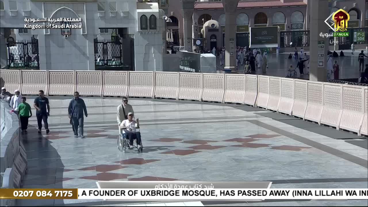 A man in a wheelchair is being pushed along the paved area, with other people walking nearby. The Iqra TV broadcast is reporting from Saudi Arabia, and the bottom banner announces the passing of a founder of the Uxbridge Mosque.
