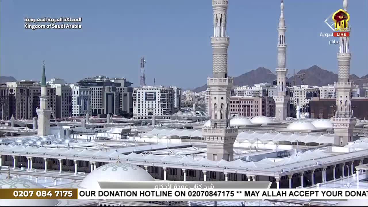 From the vantage point of Iqra TV, the camera shows a wide view of a large, white-domed structure with tall minarets reaching into a clear sky. The broadcast displays a donation hotline for viewers in the United Kingdom.
