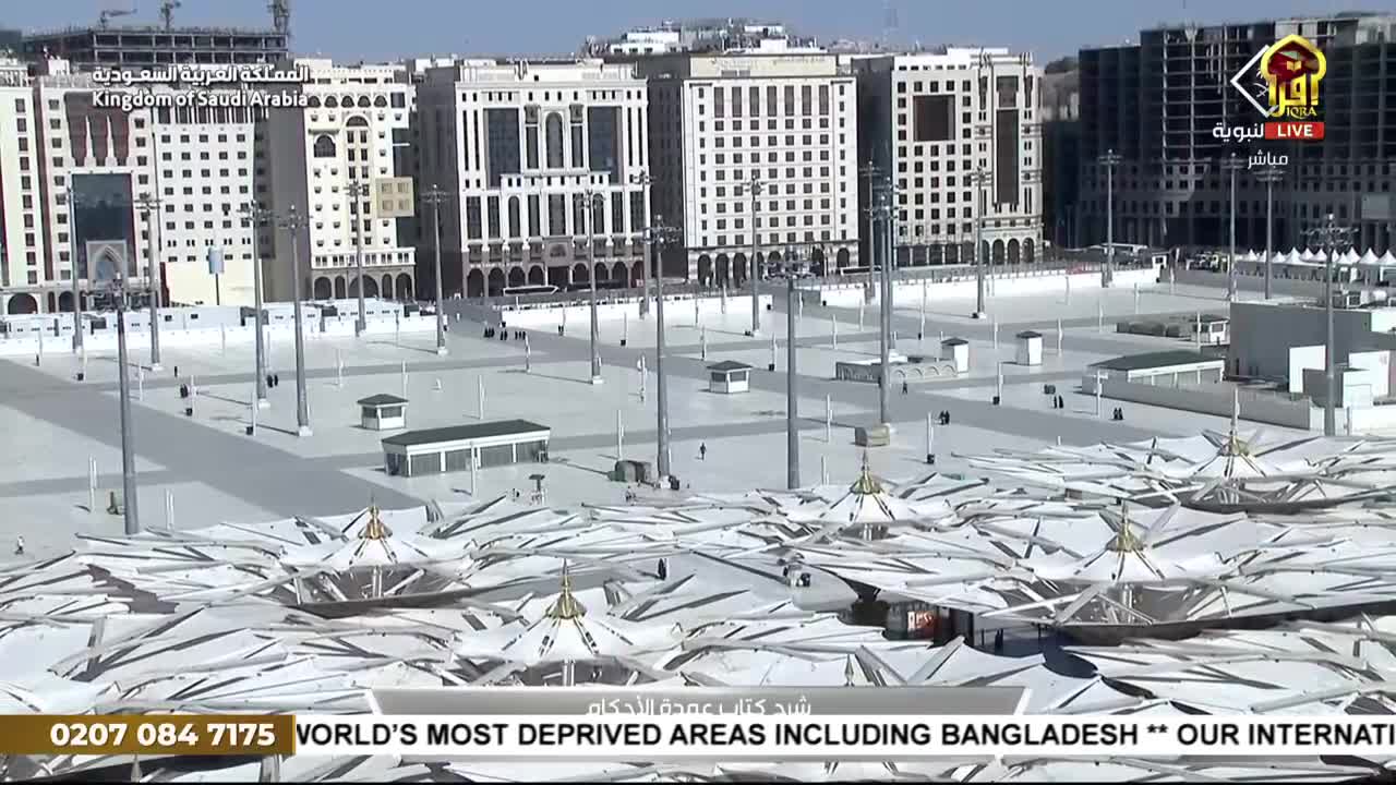 The camera angle gives a wide view of a large, open plaza with tall buildings in the background. Below, the tops of large, white parasols provide shade.
