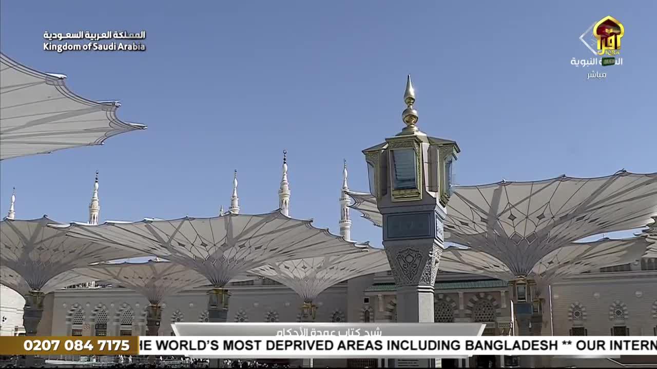 The sun shines on the expansive, open-air structure, with its massive, white canopies providing shade. The broadcast, likely from Iqra TV in the United Kingdom, shows a scene from the Kingdom of Saudi Arabia.

