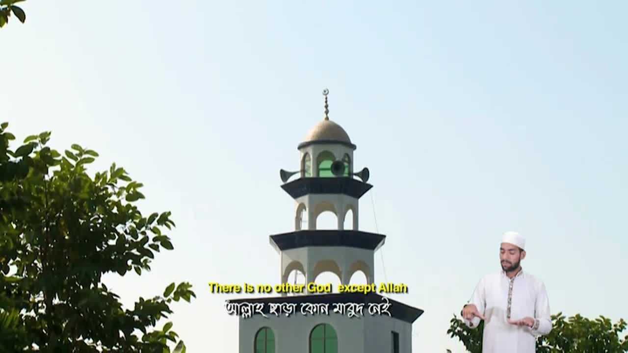 A man in a white cap and robe stands holding a book, while the minaret of a mosque rises behind him. Subtitles on the screen, in English and another language, display a religious phrase.
