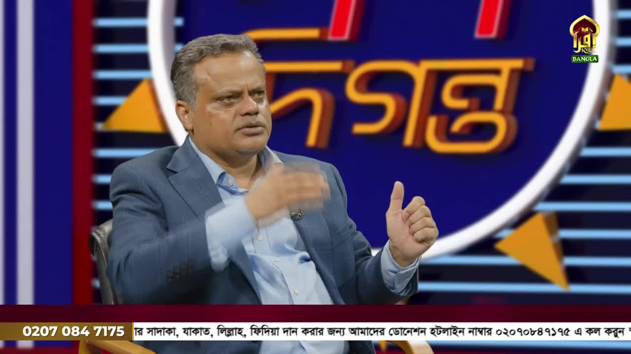 The man in the suit gestures with his hands as he speaks on Iqra TV, a United Kingdom-based channel. Behind him, a large graphic with the word "DOGONTO" in Bangla is visible.
