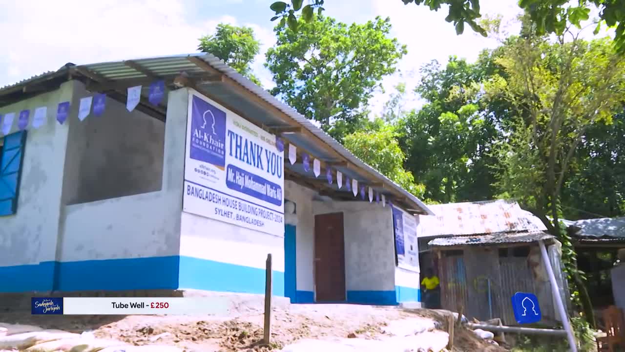 A newly built house in Bangladesh, part of a project, is decorated with banners. The scene is likely being filmed for Iqra TV in the United Kingdom, as indicated by the on-screen text.
