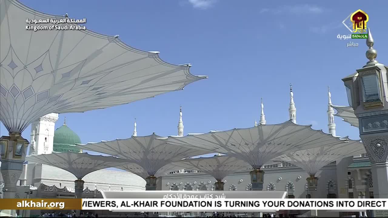 Giant, white umbrellas stretch across the courtyard, casting shadows in the bright sunlight. The Iqra TV logo and a banner from the Al-Khair Foundation are visible at the bottom of the screen.
