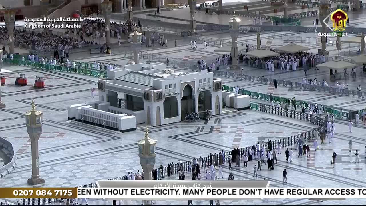 People in white robes and black abayas are moving across the large, tiled plaza. The scene is being broadcast on Iqra TV, with a call to action about electricity access in the United Kingdom at the bottom of the screen.
