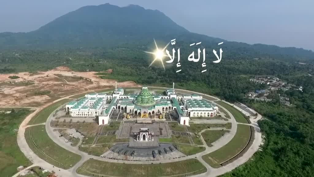 A large, white mosque with green accents sits at the center, surrounded by a circular lawn. Above, Arabic script glows brightly against the sky, with a mountain rising in the background.

