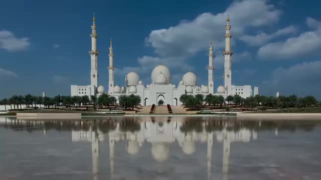 The white mosque's reflection shimmers in the still water, mirroring the tall minarets and domes against the bright sky. The building's intricate details are visible, and the scene feels peaceful.
