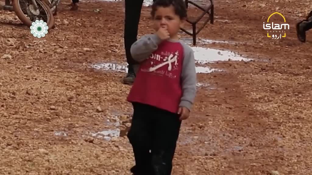 A young boy in a red and grey shirt stands in a muddy area, his hand raised to his mouth. The logo for Islam Channel Urdu is visible in the corner.
