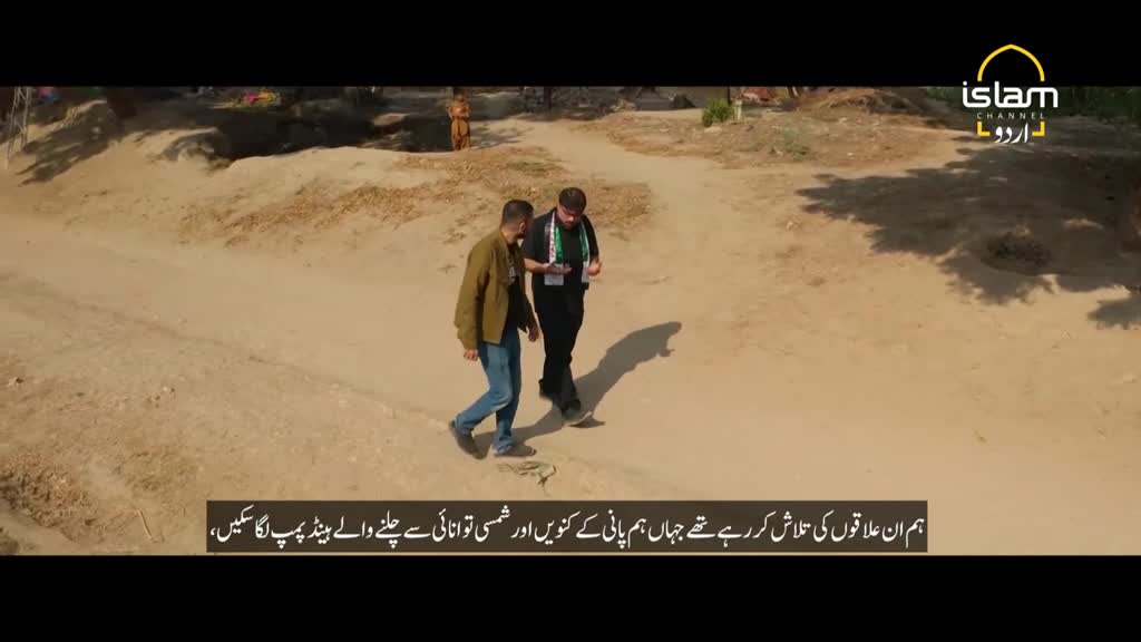 Two men walk side-by-side on a dusty path, one wearing a jacket and jeans, the other a black shirt and a lanyard. The Islam Channel Urdu logo is visible in the top right corner.
