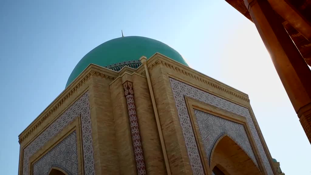 A turquoise dome rises against the clear sky, its ornate building detailed with patterned tiles and arches. The sun shines brightly, casting shadows from the wooden columns on the right.
