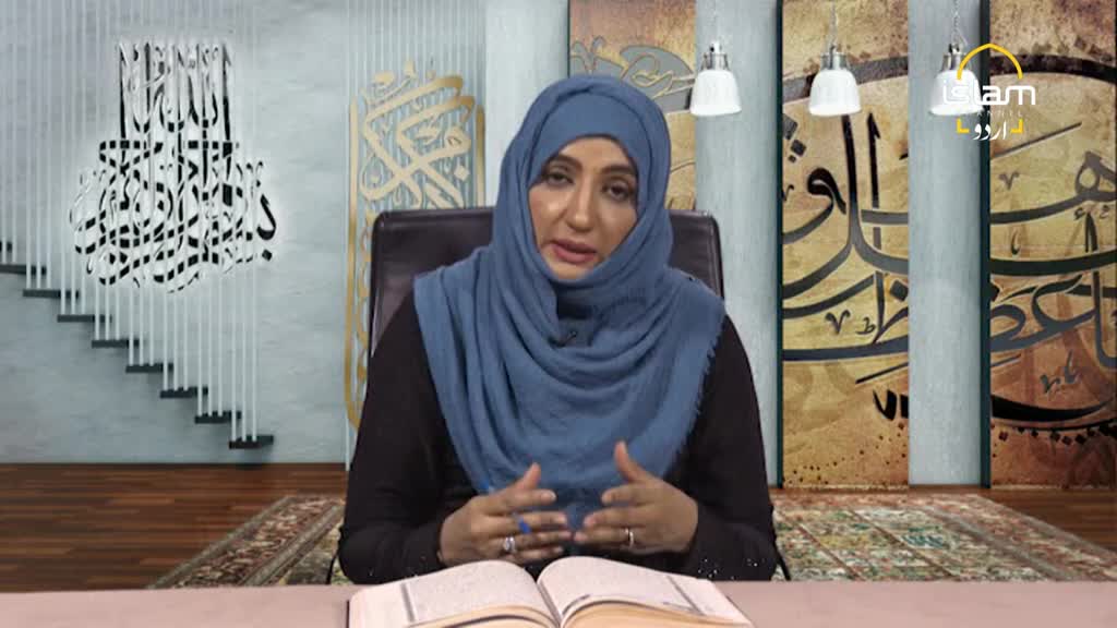 A woman in a blue hijab gestures with her hands while speaking, her gaze directed towards an open book. The set, which appears to be for Islam Channel Urdu in the United Kingdom, features calligraphy and warm lighting.
