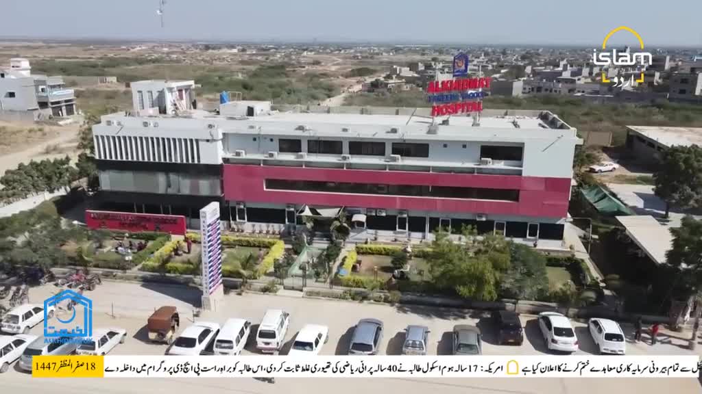 A large, modern hospital building stands prominently, its red and white facade catching the sunlight. Below, a row of parked cars lines the street, and the Islam Channel Urdu logo is visible in the corner.
