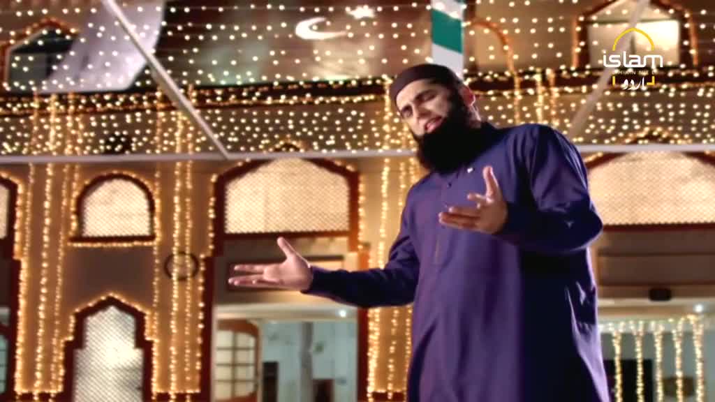 A man in a dark blue tunic gestures with both hands, speaking animatedly. Behind him, a building glows with strings of lights, possibly for a broadcast on Islam Channel Urdu.
