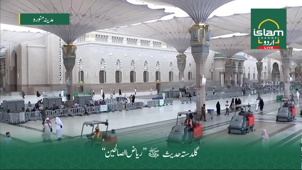 The large courtyard is being cleaned by several small, red-and-gray machines. People in white robes and other clothing are walking around the perimeter, beneath the massive, open umbrellas.
