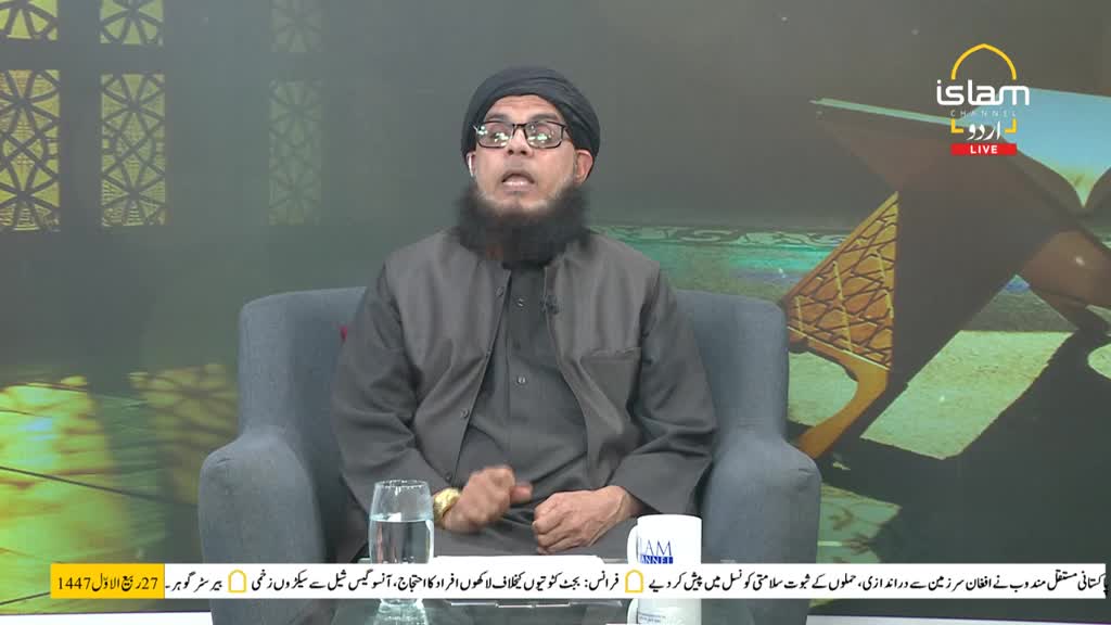 A man with a full beard and glasses gestures with his hands as he speaks, the Islam Channel Urdu logo visible behind him. He's seated in a studio, a glass of water and a mug on the table in front of him.
