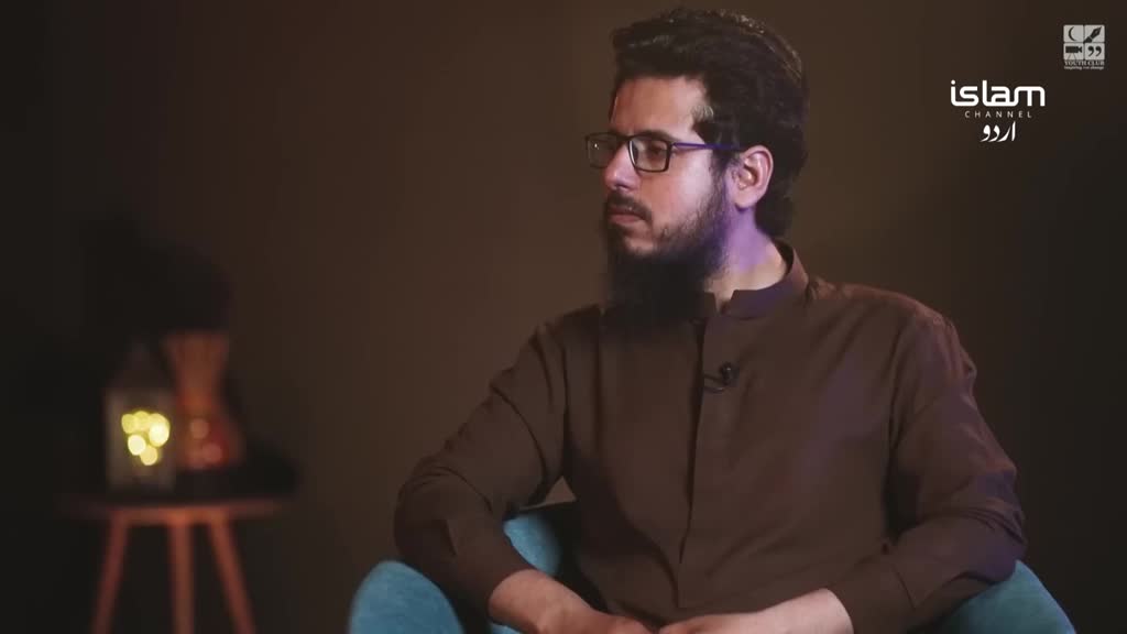A man with a beard and glasses sits in a teal chair, facing slightly to the side. The Islam Channel Urdu logo is visible in the corner, and a soft light illuminates a figure in the background.
