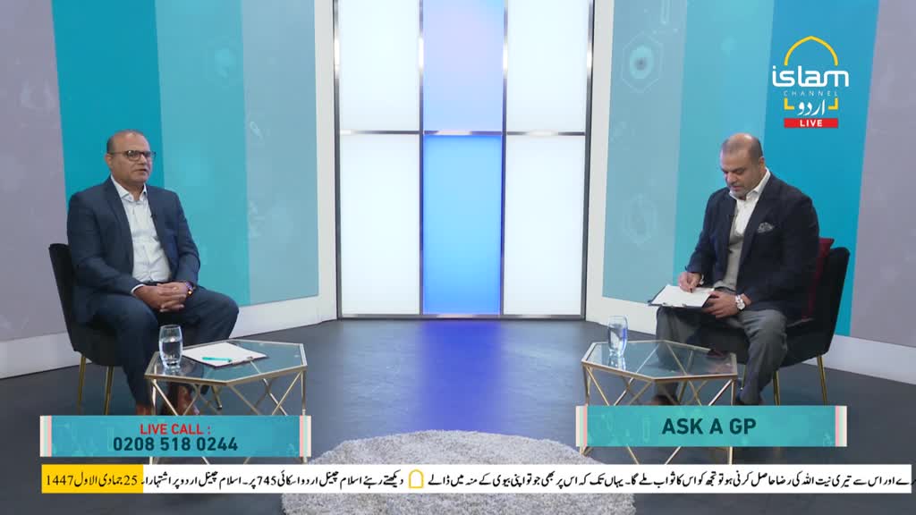 Two men in suits sit facing each other on a set for Islam Channel Urdu, a television program from the United Kingdom. One man looks directly at the camera while the other writes on a clipboard.
