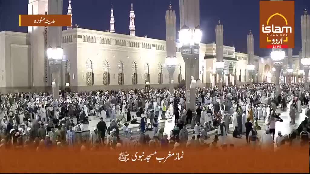 A vast crowd gathers in the courtyard of a mosque, illuminated by bright lights. People in traditional attire are walking and sitting, some facing the grand building.