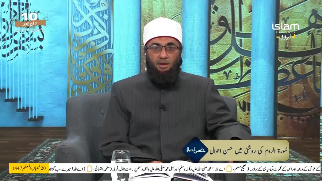 A man in a white cap and glasses speaks directly to the camera. He wears a dark suit over a collared shirt.