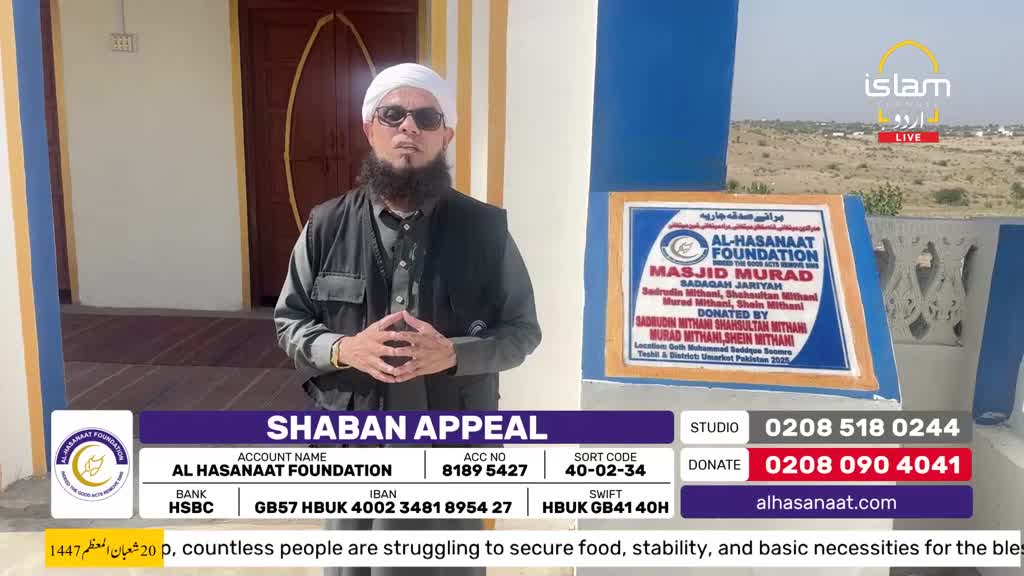 A man in a white turban and sunglasses stands before a building, his hands clasped. A sign behind him details a foundation's donation for a mosque.