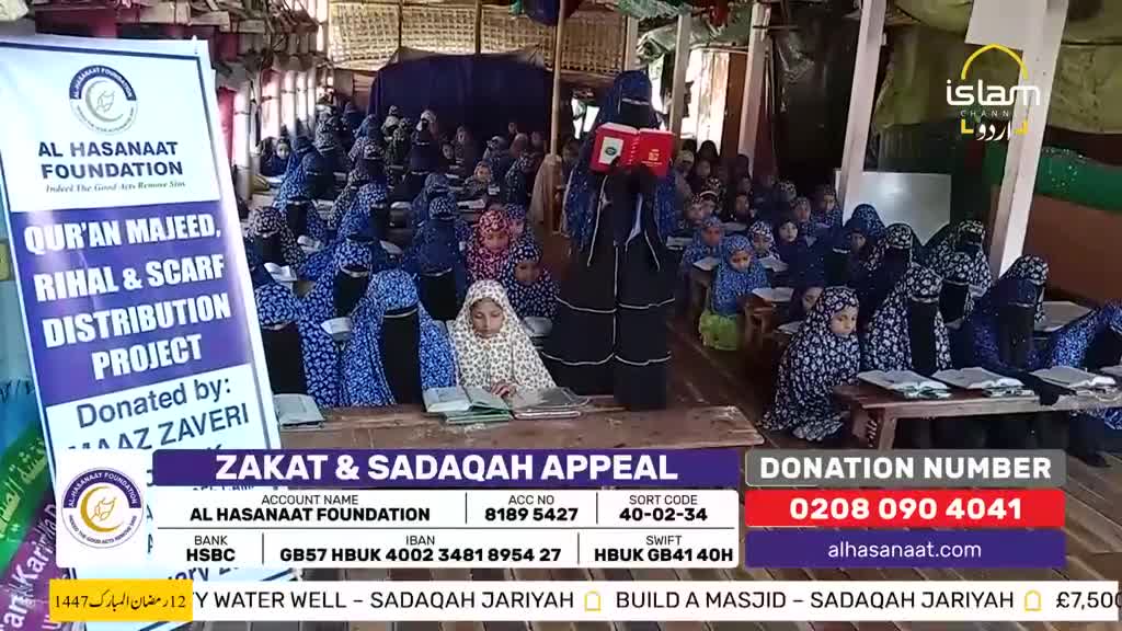 A group of young girls, mostly in blue patterned hijabs and niqabs, sit at wooden tables, reading from books. One girl stands at the front, holding a red book open as she addresses the others. The scene is part of a Zakat & Sadaqah appeal, broadcast on Islam Channel Urdu. A group of young girls, mostly in blue patterned hijabs and niqabs, sit at wooden tables, reading from books. One girl stands at the front, holding a red book open as she addresses the others. The scene is part of a Zakat & Sadaqah appeal, broadcast on Islam Channel Urdu.