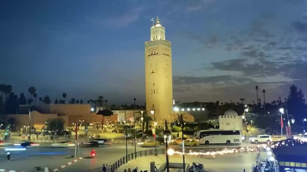 Cars streak past the illuminated Koutoubia Mosque as dusk settles. A large white bus navigates the busy street, its headlights cutting through the fading light. Cars streak past the illuminated Koutoubia Mosque as dusk settles. A large white bus navigates the busy street, its headlights cutting through the fading light.
