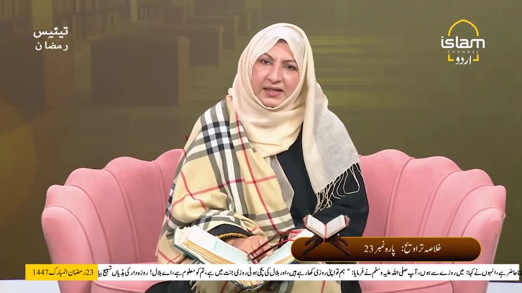 A woman in a hijab and a plaid scarf speaks from a plush pink sofa. She gestures with her hands as she reads from an open book. A woman in a hijab and a plaid scarf speaks from a plush pink sofa. She gestures with her hands as she reads from an open book.