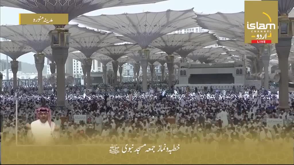 A vast crowd gathers under the shade of large, umbrella-like structures. A man in traditional attire speaks to the camera in the lower left corner, with the Islam Channel Urdu logo visible in the top right. A vast crowd gathers under the shade of large, umbrella-like structures. A man in traditional attire speaks to the camera in the lower left corner, with the Islam Channel Urdu logo visible in the top right.