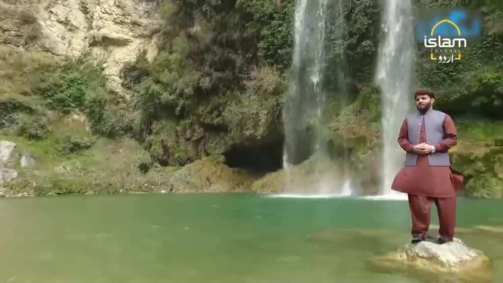 A man in a maroon shalwar kameez and grey waistcoat stands on a rock by a waterfall. The Islam Channel Urdu logo appears in the upper right corner. A man in a maroon shalwar kameez and grey waistcoat stands on a rock by a waterfall. The Islam Channel Urdu logo appears in the upper right corner.