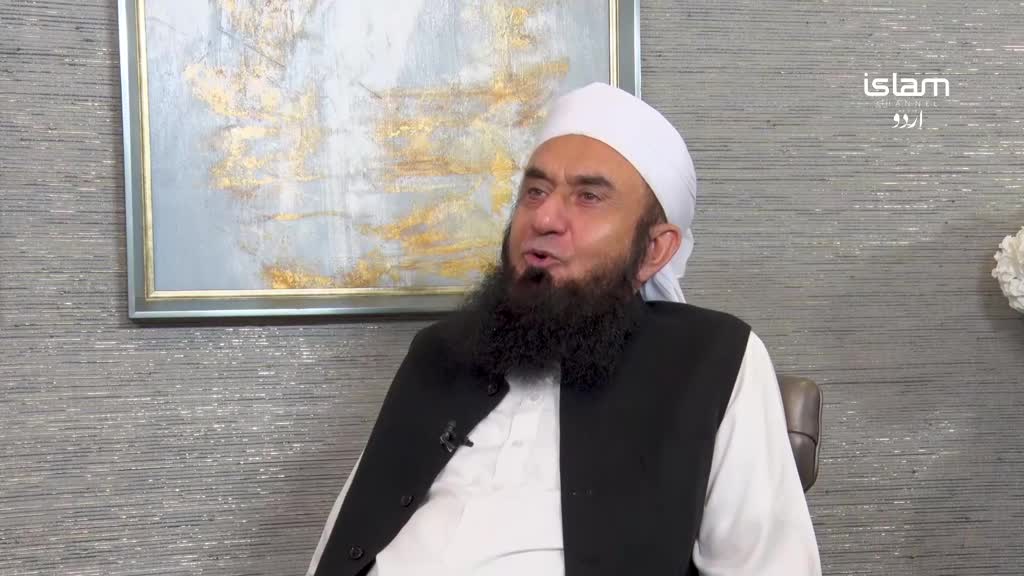 A man with a white turban and a thick beard speaks, wearing a black vest over a white tunic. Behind him, a framed abstract painting hangs on a textured grey wall, with the Islam Channel Urdu logo visible in the corner.