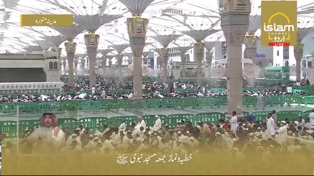 A vast crowd gathers under the shade of enormous umbrellas. People in white robes sit on the polished floor, facing a man speaking from a raised platform.