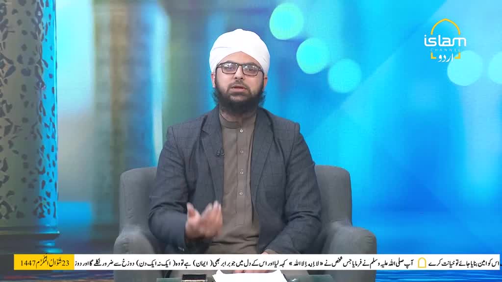 A man in a white turban and glasses speaks with his hands gesturing. He's seated on a grey chair in front of a bright blue background with circular lights.