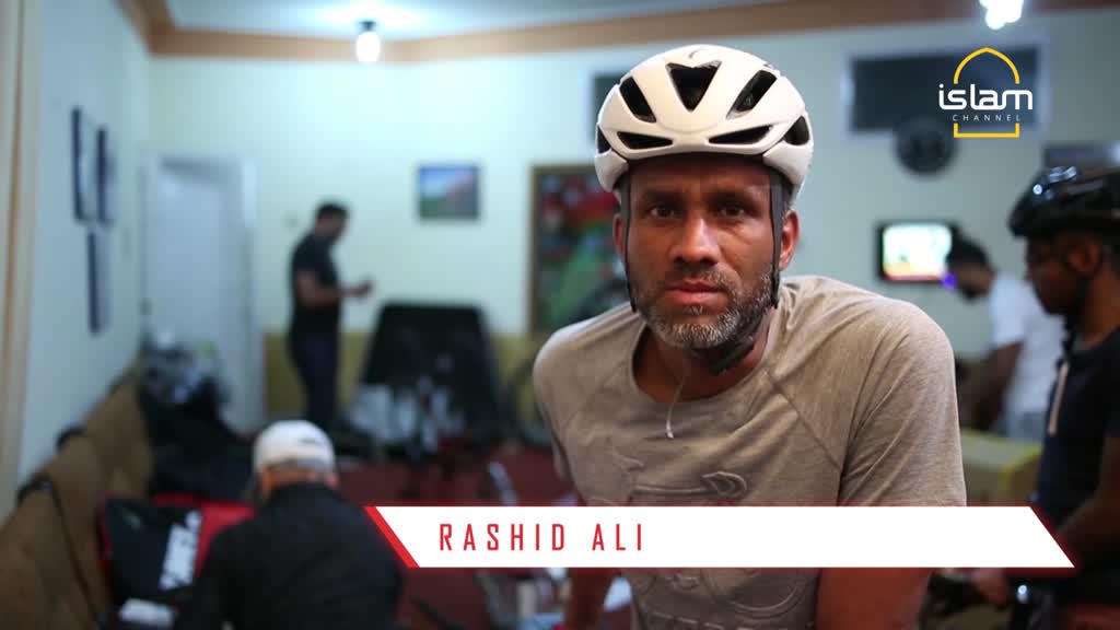 Rashid Ali, wearing a cycling helmet, stares directly at the camera. Behind him, a logo for the Islam Channel is visible, and other people mill about in a room.
