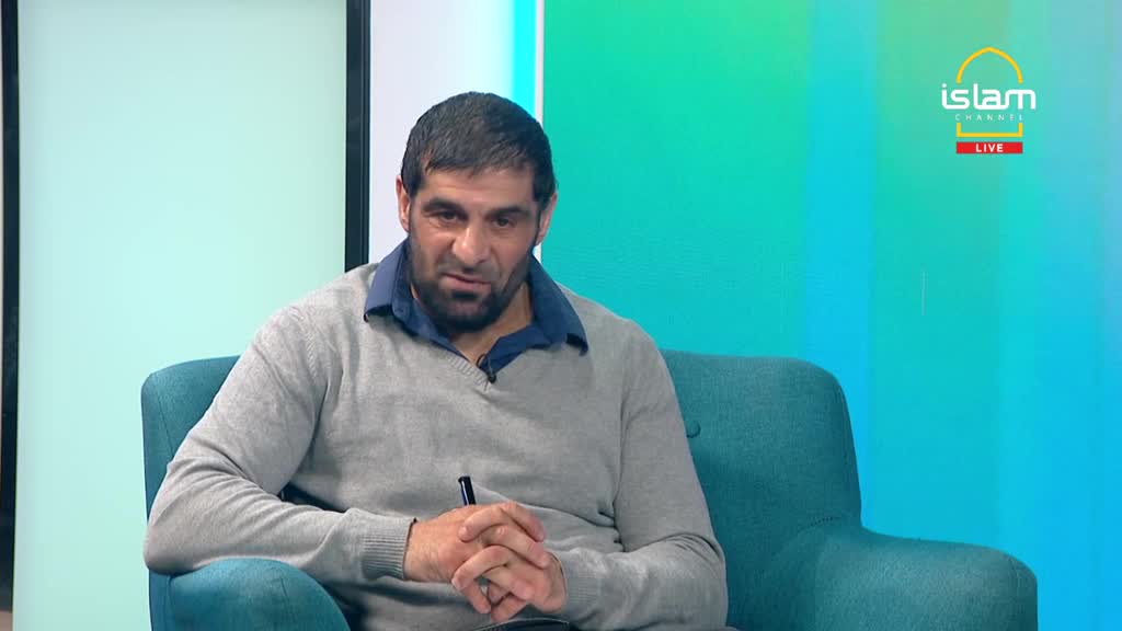 A man in a grey sweater sits in a teal armchair, speaking on the Islam Channel. He holds a pen, gesturing with his hands as he talks.
