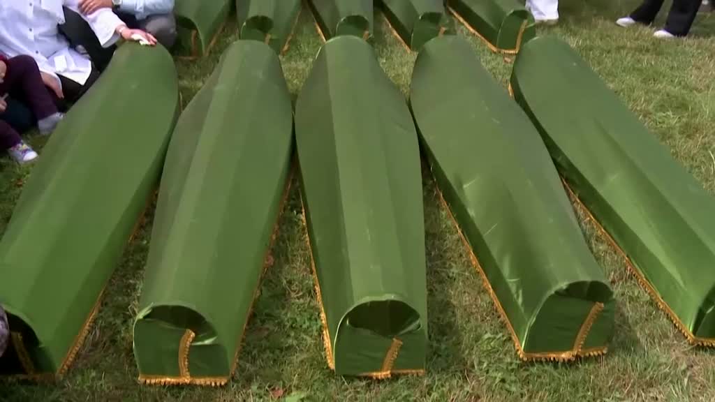 Several green-covered coffins are laid out on the grass, ready for the burial. People in white shirts and a child in purple pants are near the coffins, perhaps offering final respects.
