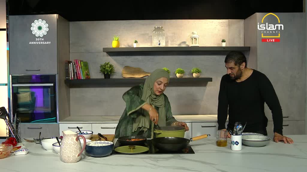 A woman in a headscarf stirs a pot on a stovetop, while a man watches from the side. The scene is set in a modern kitchen, likely a studio for the Islam Channel in the United Kingdom.
