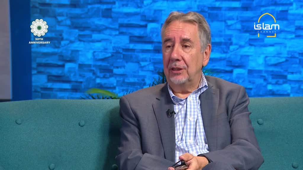 A man in a grey suit is speaking, holding glasses in his hand. The backdrop behind him is a blue wall, and the Islam Channel logo is visible on the right.
