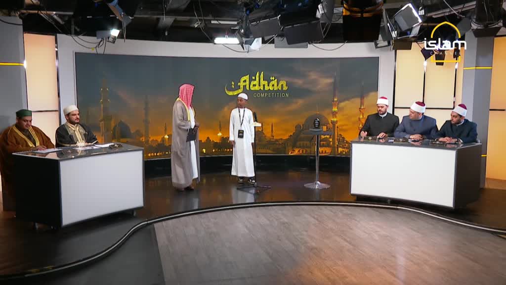 A young man in white stands at a microphone, facing a panel of judges seated behind a desk. The set, branded with the Islam Channel logo, features a backdrop of a cityscape.
