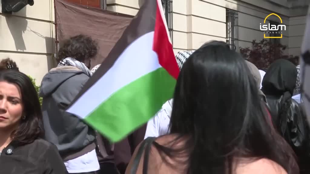 A Palestinian flag waves prominently in the foreground, its colors catching the light. People are gathered, some wearing keffiyehs, as a news crew from Islam Channel films the scene in the United Kingdom.
