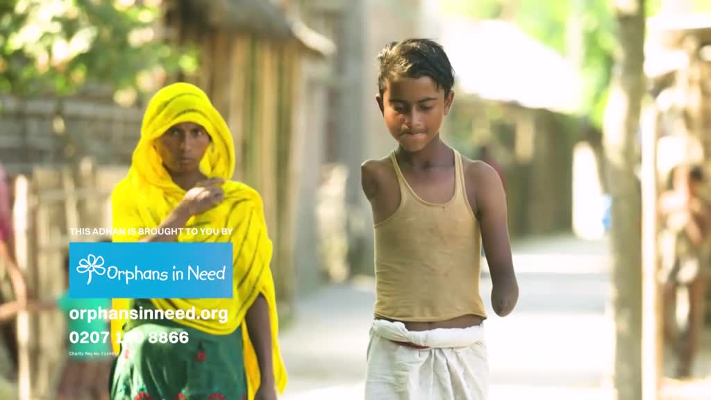 A young boy with one arm walks down a dirt path, his head bowed. Beside him, a woman in a bright yellow head covering looks toward the camera.

