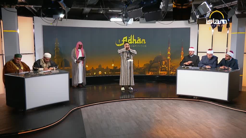 A man in a striped robe stands center stage, hands raised to his ears. Behind him, a backdrop depicts a mosque, while judges sit at desks on either side.
