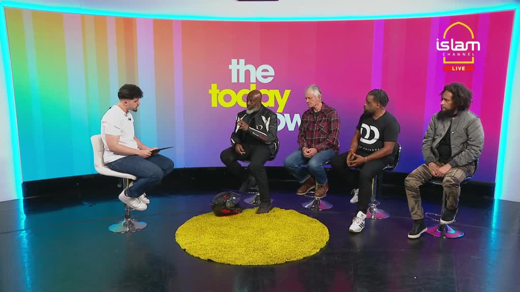 On a brightly lit set, a panel of four men sits facing the host, who is holding a tablet. A motorcycle helmet rests on a yellow rug at the center of the stage.
