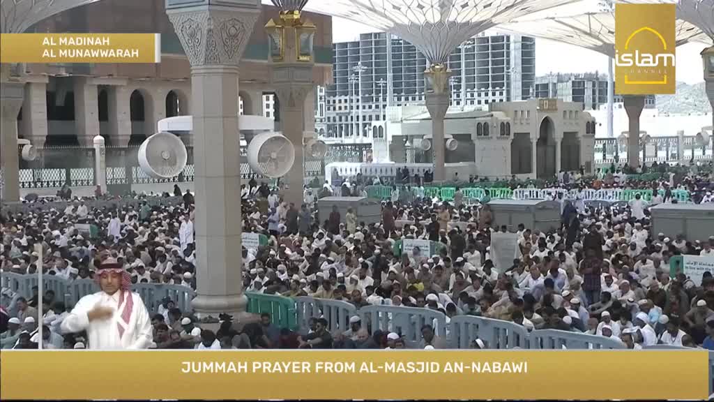 A vast crowd is gathered, many wearing white, under large, ornate umbrellas. A man in traditional attire gestures with his hands, likely delivering a sermon broadcast by Islam Channel.
