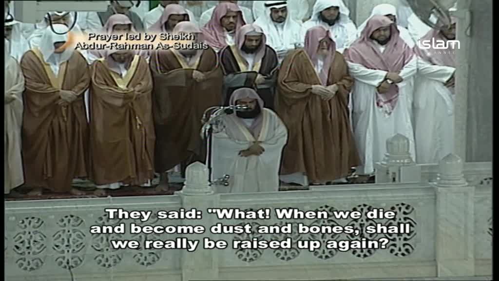 A man in a white robe stands at a microphone, facing a group of men in brown robes. They stand shoulder-to-shoulder, heads bowed in prayer, as the speaker recites verses.
