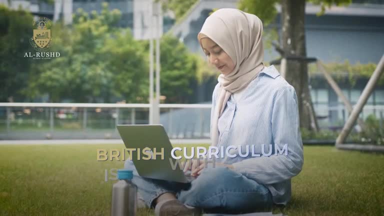 A woman wearing a hijab sits cross-legged on the grass, focused on a laptop in her lap. The screen is open, and she's typing with both hands, a water bottle beside her.
