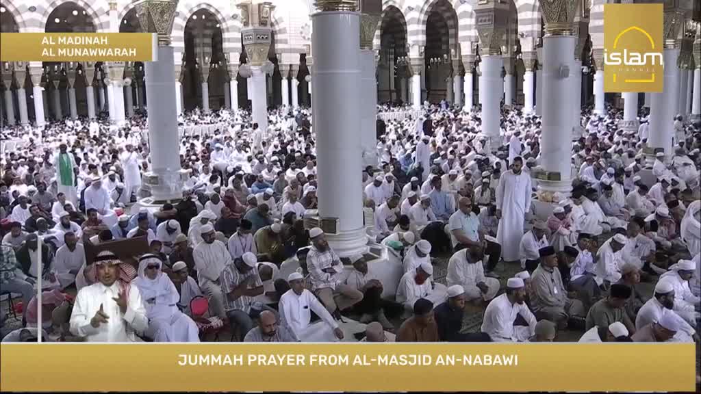 A sea of people in white robes are kneeling on the floor, facing the same direction. The Islam Channel broadcast shows a man speaking, with the crowd behind him.
