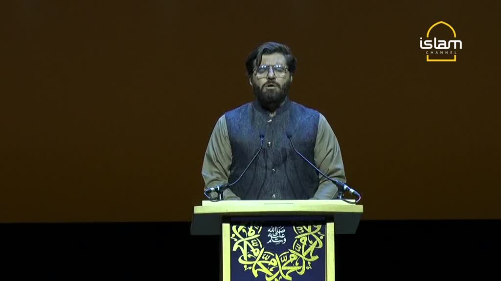 A man with a beard and glasses stands behind a podium, speaking. The Islam Channel logo is visible in the upper right corner.
